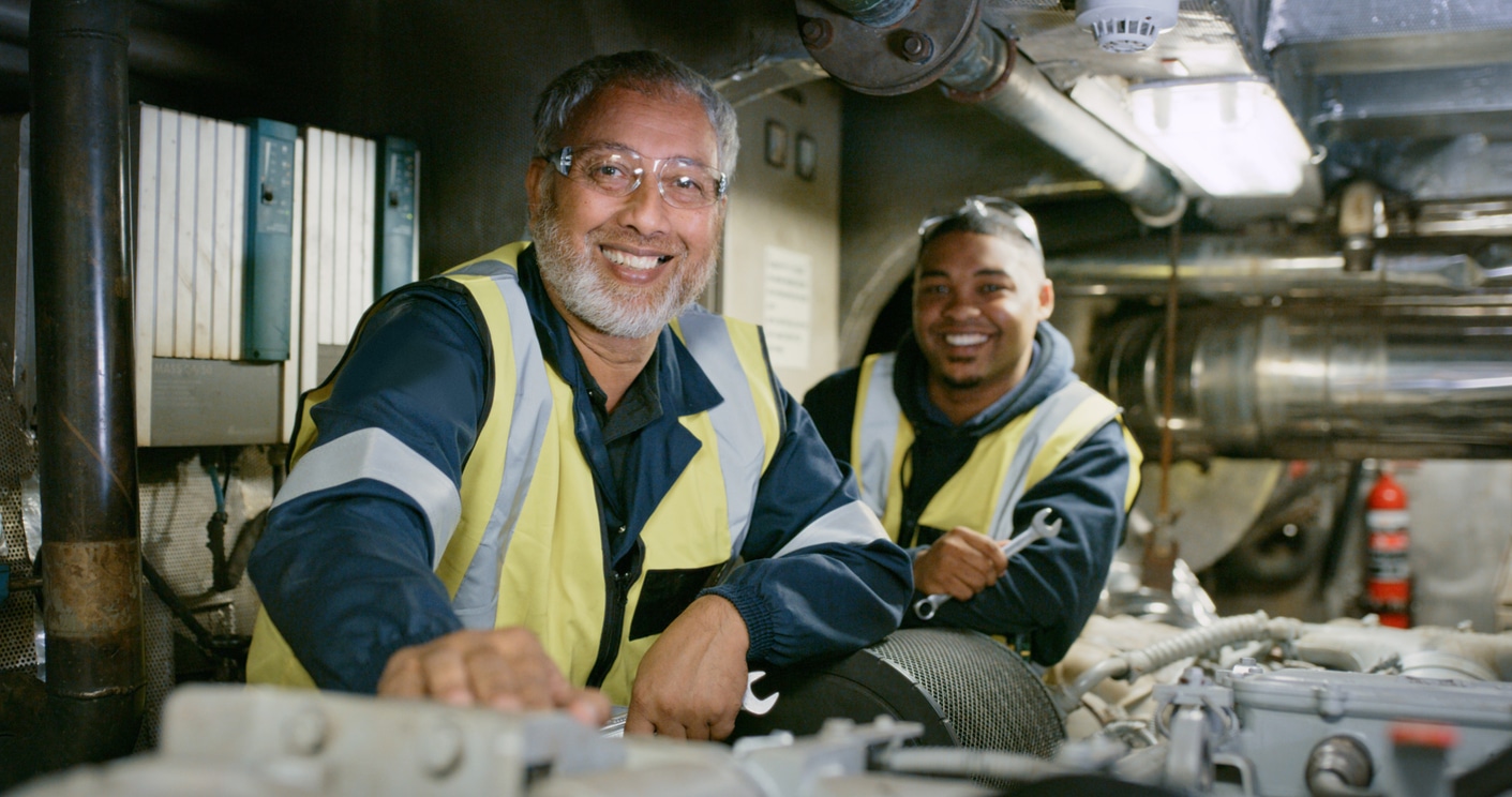 Two diesel technicians pause to smile while working in mechanics shop