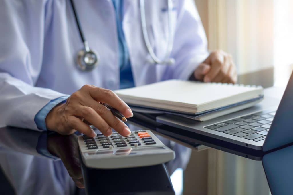 Physician with stethoscope using calculator and working on computer and notebook